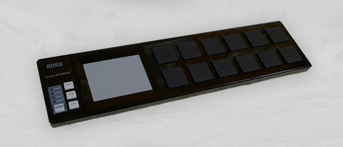 controlador Korg nano pad color negro sobre un fondo blanco, a su izquierda tiene 12 patches de goma y una pequeña pantalla que se controla con el dedo deslizandolo arriba y abajo o izquierda y derecha. También hay tres botones con funciones especificas más otro botón para cambiar la escena del controlador