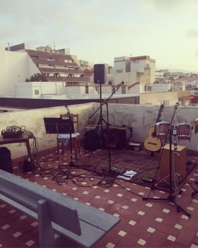 Azotea con equipo e instrumentos musicales dispuesta para comenzar un concierto. Se muestra una guitarra, un amplificador de bajo, unos bongos, un cajón flamenco, tres pies de micrófono con los micrófonos. Al fondo hay un edificio y se muestra el cielo.