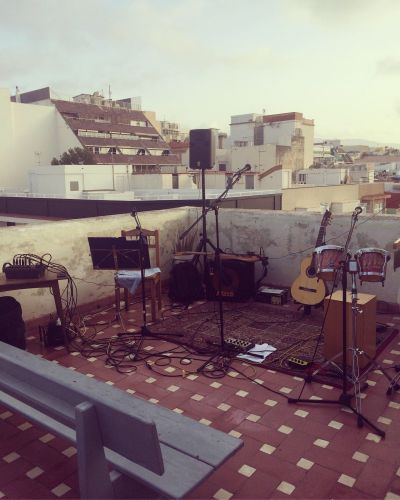 Azotea con equipo e instrumentos musicales dispuesta para comenzar un concierto. Se muestra una guitarra, un amplificador de bajo, unos bongos, un cajón flamenco, tres pies de micrófono con los micrófonos. Al fondo hay un edificio y se muestra el cielo.